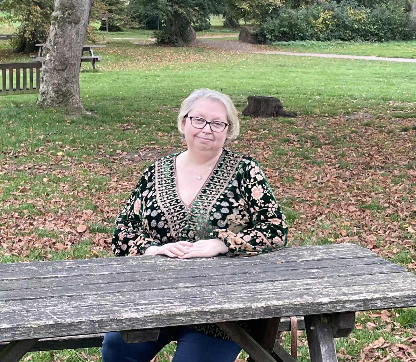 Lisa Donahue photo Lisa sits at a wooden table in a park surrounded by autumn leaves.
